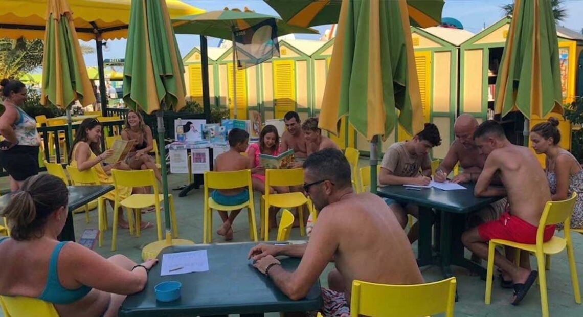 Laboratorio didattico all'aperto per bambini e famiglie presso uno stabilimento balneare a Pesaro, con tavoli gialli, ombrelloni e cabine sullo sfondo.
