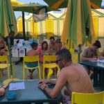 Laboratorio didattico all'aperto per bambini e famiglie presso uno stabilimento balneare a Pesaro, con tavoli gialli, ombrelloni e cabine sullo sfondo.