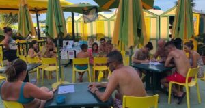 Laboratorio didattico all'aperto per bambini e famiglie presso uno stabilimento balneare a Pesaro, con tavoli gialli, ombrelloni e cabine sullo sfondo.