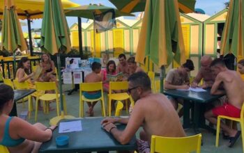 Laboratorio didattico all'aperto per bambini e famiglie presso uno stabilimento balneare a Pesaro, con tavoli gialli, ombrelloni e cabine sullo sfondo.