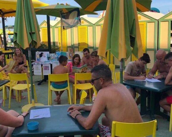 Laboratorio didattico all'aperto per bambini e famiglie presso uno stabilimento balneare a Pesaro, con tavoli gialli, ombrelloni e cabine sullo sfondo.