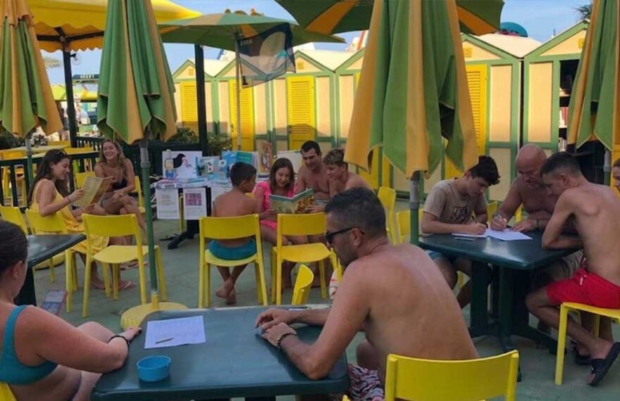 Laboratorio didattico all'aperto per bambini e famiglie presso uno stabilimento balneare a Pesaro, con tavoli gialli, ombrelloni e cabine sullo sfondo.
