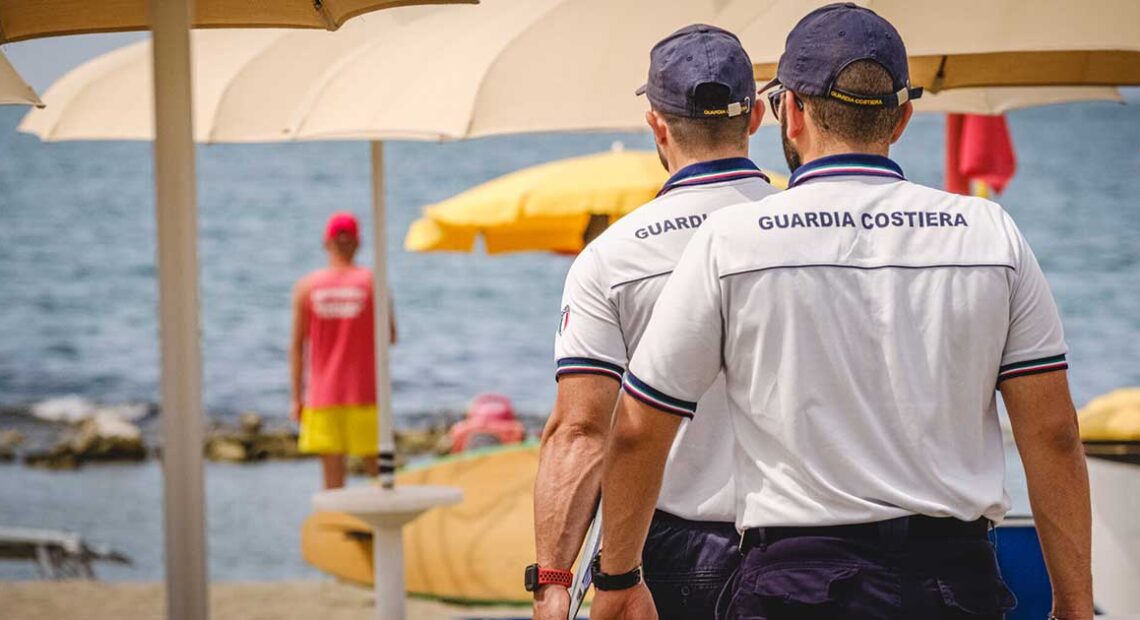 Militari della Guardia Costiera in divisa effettuano controlli tra gli ombrelloni in uno stabilimento balneare.