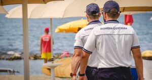Militari della Guardia Costiera in divisa effettuano controlli tra gli ombrelloni in uno stabilimento balneare.