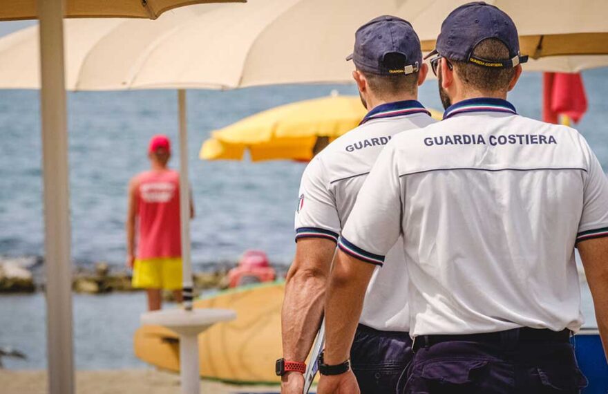 Militari della Guardia Costiera in divisa effettuano controlli tra gli ombrelloni in uno stabilimento balneare.
