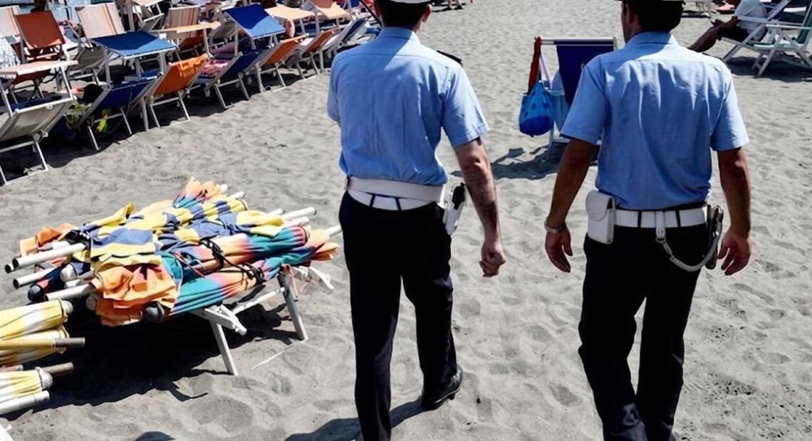 Agenti della Polizia Municipale in pattugliamento sulla spiaggia libera accanto a una pila di ombrelloni e lettini sequestrati.