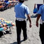 Agenti della Polizia Municipale in pattugliamento sulla spiaggia libera accanto a una pila di ombrelloni e lettini sequestrati.