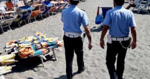 Agenti della Polizia Municipale in pattugliamento sulla spiaggia libera accanto a una pila di ombrelloni e lettini sequestrati.