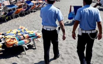 Agenti della Polizia Municipale in pattugliamento sulla spiaggia libera accanto a una pila di ombrelloni e lettini sequestrati.