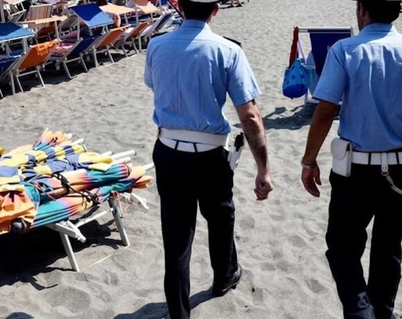 Agenti della Polizia Municipale in pattugliamento sulla spiaggia libera accanto a una pila di ombrelloni e lettini sequestrati.