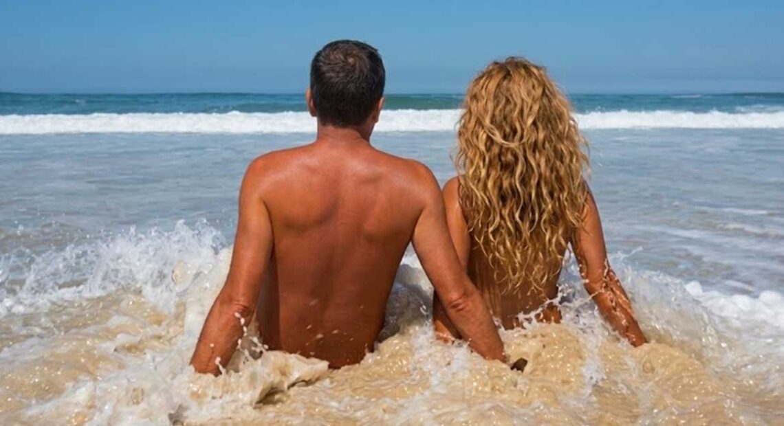 Coppia di spalle seduta sulla battigia bagnata dalle onde del mare in una spiaggia naturista