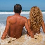 Coppia di spalle seduta sulla battigia bagnata dalle onde del mare in una spiaggia naturista