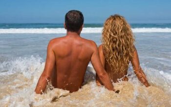 Coppia di spalle seduta sulla battigia bagnata dalle onde del mare in una spiaggia naturista