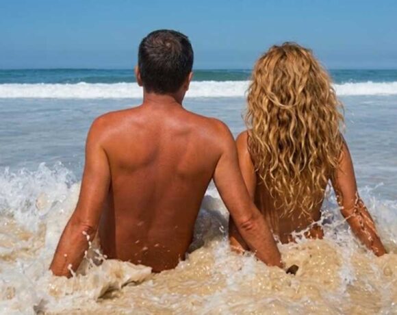 Coppia di spalle seduta sulla battigia bagnata dalle onde del mare in una spiaggia naturista