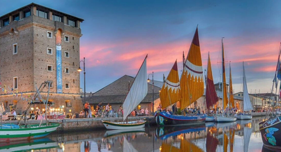 Veduta suggestiva del Porto Canale di Cervia al tramonto con la Torre San Michele illuminata e le storiche barche da pesca con vele al terzo spiegate.