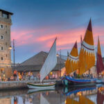 Veduta suggestiva del Porto Canale di Cervia al tramonto con la Torre San Michele illuminata e le storiche barche da pesca con vele al terzo spiegate.