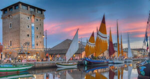 Veduta suggestiva del Porto Canale di Cervia al tramonto con la Torre San Michele illuminata e le storiche barche da pesca con vele al terzo spiegate.