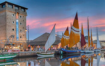 Veduta suggestiva del Porto Canale di Cervia al tramonto con la Torre San Michele illuminata e le storiche barche da pesca con vele al terzo spiegate.
