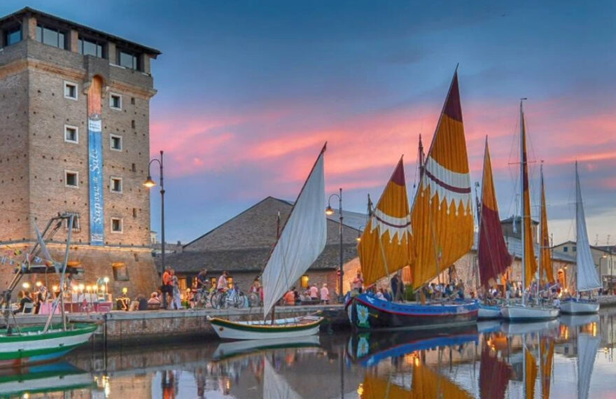 Veduta suggestiva del Porto Canale di Cervia al tramonto con la Torre San Michele illuminata e le storiche barche da pesca con vele al terzo spiegate.