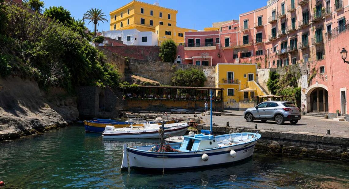 Scorcio del Porto Romano di Ventotene con barche da pesca ormeggiate e palazzi storici colorati sullo sfondo sotto un cielo azzurro.