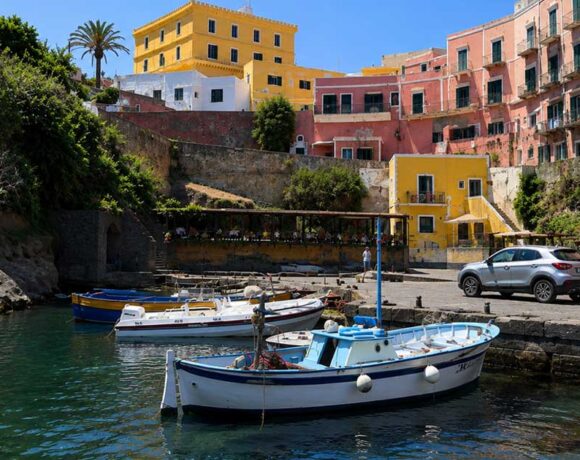 Scorcio del Porto Romano di Ventotene con barche da pesca ormeggiate e palazzi storici colorati sullo sfondo sotto un cielo azzurro.