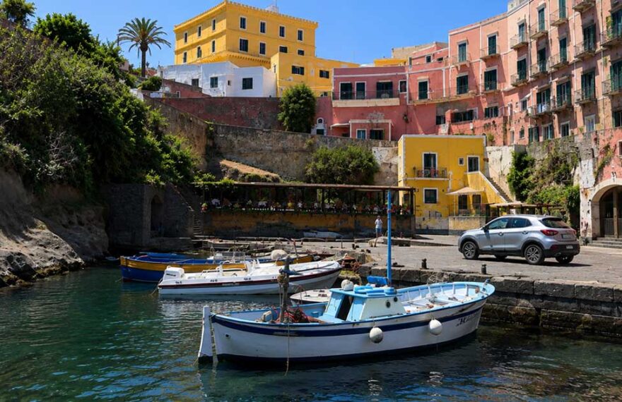 Scorcio del Porto Romano di Ventotene con barche da pesca ormeggiate e palazzi storici colorati sullo sfondo sotto un cielo azzurro.