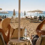Vista posteriore di una coppia seduta su lettini sotto un ombrellone beige su una spiaggia affollata in una giornata soleggiata. La donna usa un ventaglio.