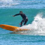 Un surfista in muta nera in equilibrio su una tavola arancione tra le onde del mare ligure.
