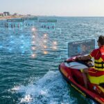 Un bagnino in divisa rossa su una moto d'acqua diretta verso il largo, con elementi grafici digitali di intelligenza artificiale (immagine generata con IA)