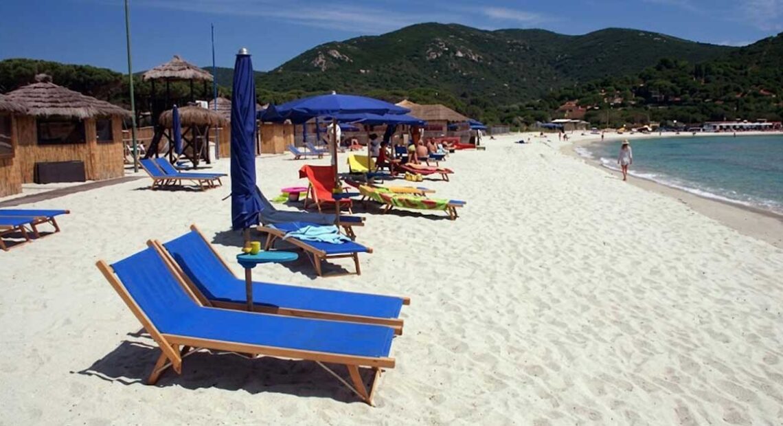 Spiaggia dell'Isola d'Elba con sabbia bianca, lettini blu e ombrelloni con colline sullo sfondo