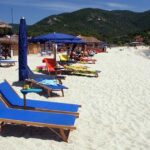 Spiaggia dell'Isola d'Elba con sabbia bianca, lettini blu e ombrelloni con colline sullo sfondo