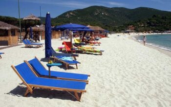 Spiaggia dell'Isola d'Elba con sabbia bianca, lettini blu e ombrelloni con colline sullo sfondo