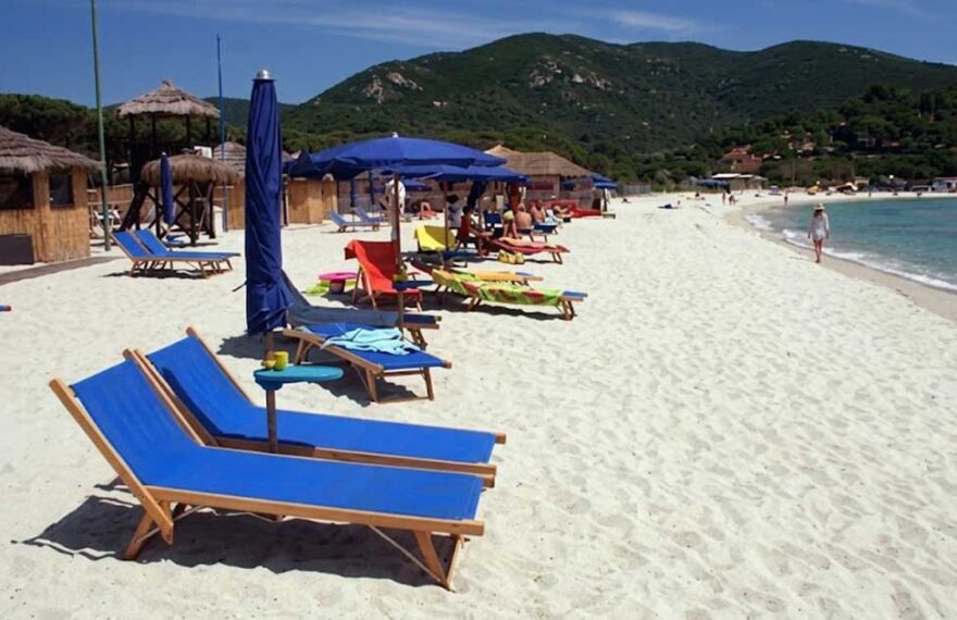 Spiaggia dell'Isola d'Elba con sabbia bianca, lettini blu e ombrelloni con colline sullo sfondo