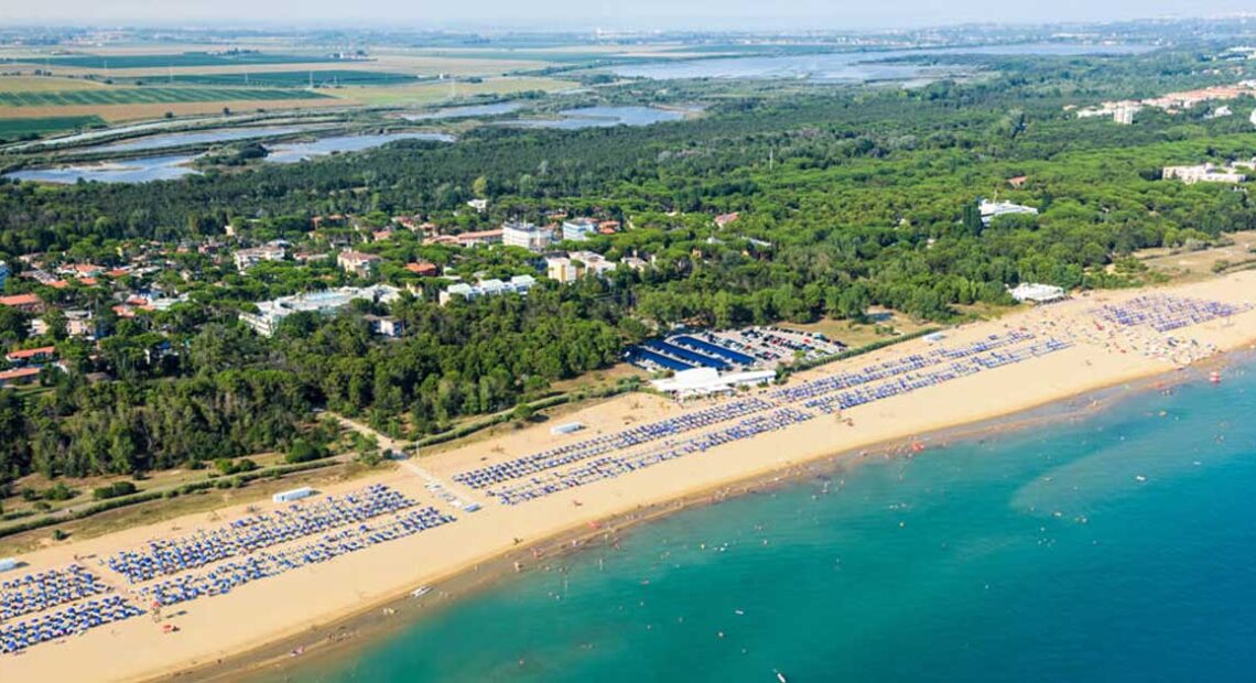 Fotografia aerea dell'ampio litorale sabbioso di Bibione, popolato da stabilimenti balneari con ombrelloni e bagnanti, che confina con una fitta pineta e un insediamento urbano sullo sfondo.