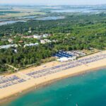 Fotografia aerea dell'ampio litorale sabbioso di Bibione, popolato da stabilimenti balneari con ombrelloni e bagnanti, che confina con una fitta pineta e un insediamento urbano sullo sfondo.
