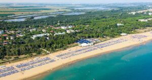 Fotografia aerea dell'ampio litorale sabbioso di Bibione, popolato da stabilimenti balneari con ombrelloni e bagnanti, che confina con una fitta pineta e un insediamento urbano sullo sfondo.