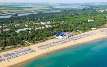 Fotografia aerea dell'ampio litorale sabbioso di Bibione, popolato da stabilimenti balneari con ombrelloni e bagnanti, che confina con una fitta pineta e un insediamento urbano sullo sfondo.