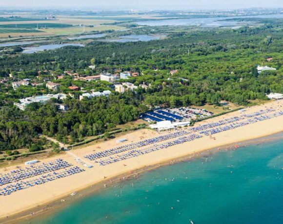 Fotografia aerea dell'ampio litorale sabbioso di Bibione, popolato da stabilimenti balneari con ombrelloni e bagnanti, che confina con una fitta pineta e un insediamento urbano sullo sfondo.