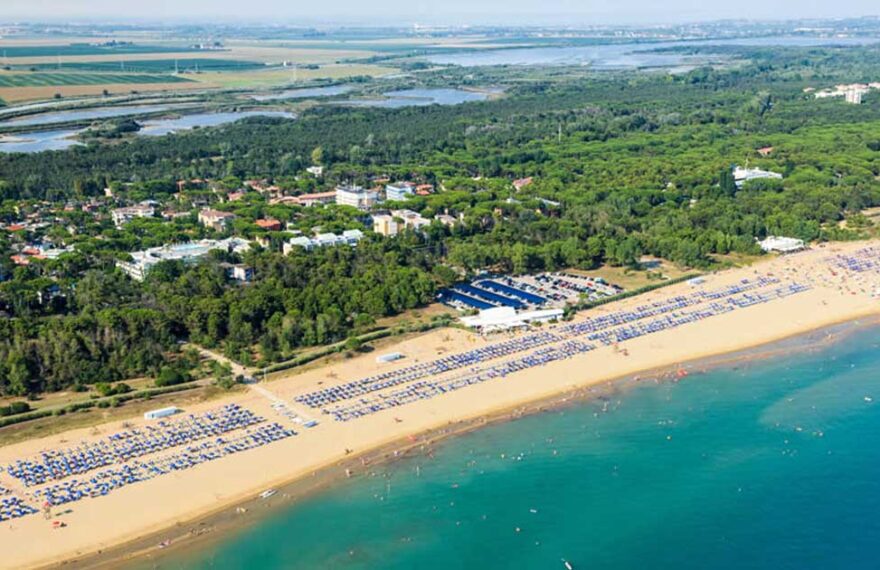 Fotografia aerea dell'ampio litorale sabbioso di Bibione, popolato da stabilimenti balneari con ombrelloni e bagnanti, che confina con una fitta pineta e un insediamento urbano sullo sfondo.