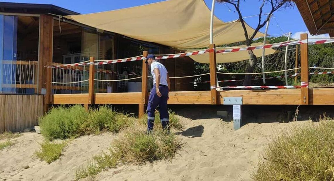 Militare della Guardia Costiera accanto a una veranda in legno sigillata su una duna di sabbia.
