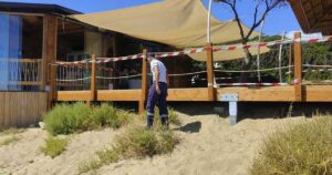Militare della Guardia Costiera accanto a una veranda in legno sigillata su una duna di sabbia.