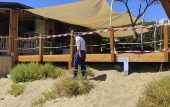 Militare della Guardia Costiera accanto a una veranda in legno sigillata su una duna di sabbia.