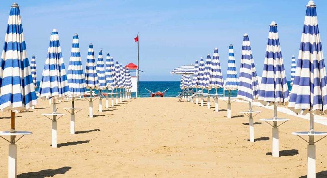 File ordinate di ombrelloni chiusi a strisce bianche e blu sulla spiaggia deserta di Rimini con torretta di salvataggio sullo sfondo