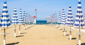 File ordinate di ombrelloni chiusi a strisce bianche e blu sulla spiaggia deserta di Rimini con torretta di salvataggio sullo sfondo