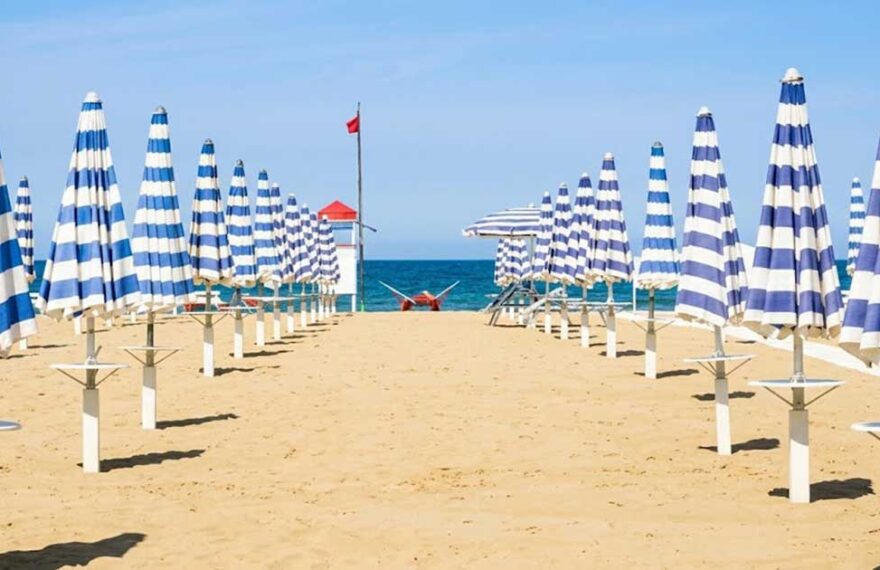 File ordinate di ombrelloni chiusi a strisce bianche e blu sulla spiaggia deserta di Rimini con torretta di salvataggio sullo sfondo
