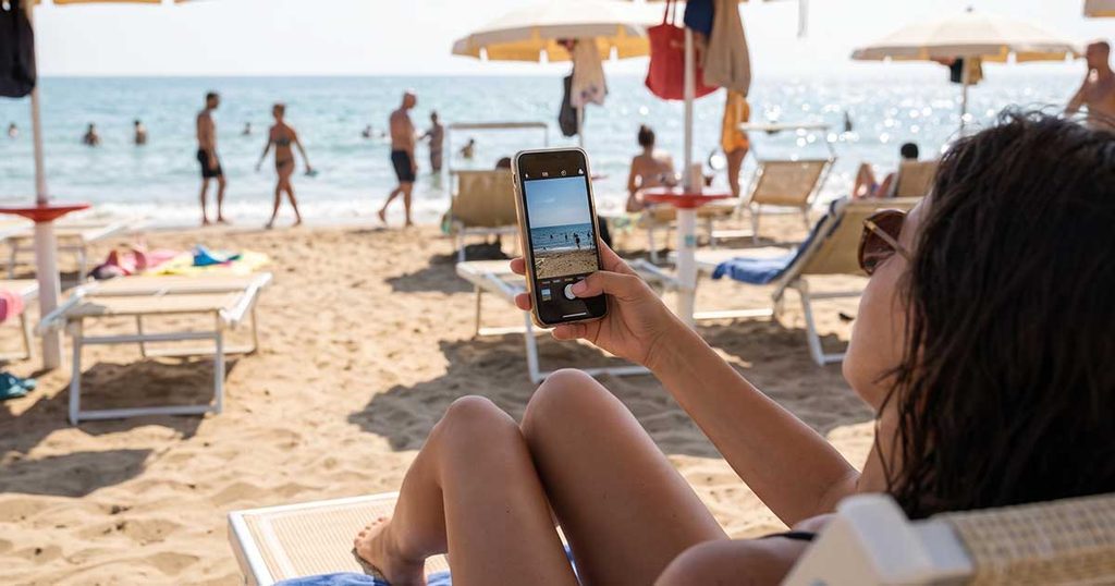 Una bagnante seduta su un lettino in spiaggia che scatta o condivide una foto del mare con lo smartphone sotto l'ombrellone.