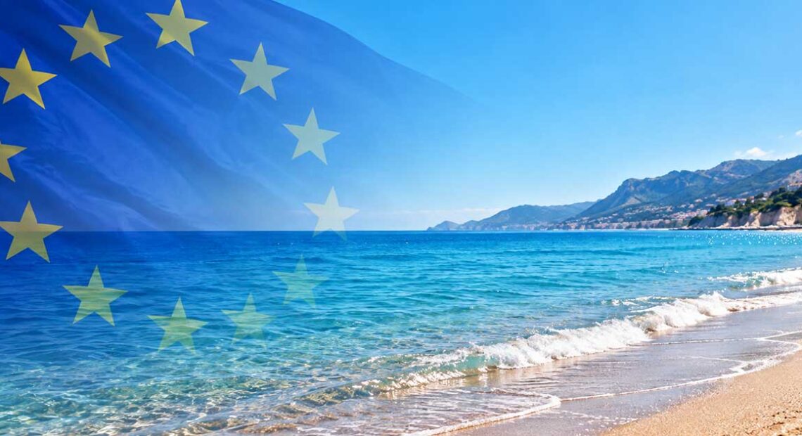 Bandiera dell'Unione Europea in trasparenza su una spiaggia mediterranea con mare limpido e costa montuosa per la decisione della Commissione UE sulle concessioni balneari.
