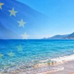 Bandiera dell'Unione Europea in trasparenza su una spiaggia mediterranea con mare limpido e costa montuosa per la decisione della Commissione UE sulle concessioni balneari.