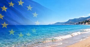Bandiera dell'Unione Europea in trasparenza su una spiaggia mediterranea con mare limpido e costa montuosa per la decisione della Commissione UE sulle concessioni balneari.