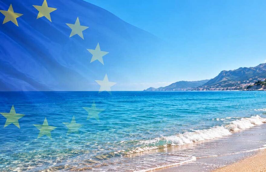 Bandiera dell'Unione Europea in trasparenza su una spiaggia mediterranea con mare limpido e costa montuosa per la decisione della Commissione UE sulle concessioni balneari.